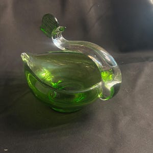 May include: A green glass bowl shaped like a bird, featuring a deep emerald green basin and a clear, curved glass neck and head that extends upward from the side, creating a smooth, continuous silhouette with a distinct avian form.