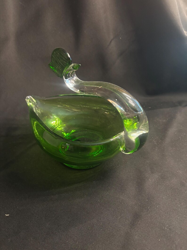 May include: A green glass bowl shaped like a bird, featuring a deep emerald green basin and a clear, curved glass neck and head that extends upward from the side, creating a smooth, continuous silhouette with a distinct avian form.