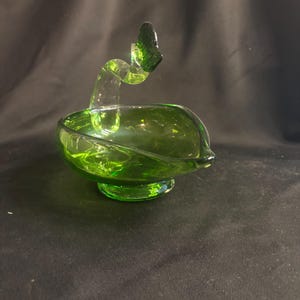 May include: A translucent emerald green glass bowl featuring a curved, swan-like neck handle that extends upward from the rim. The vessel rests on a circular pedestal base and has a smooth, polished finish with a pointed spout on one side.