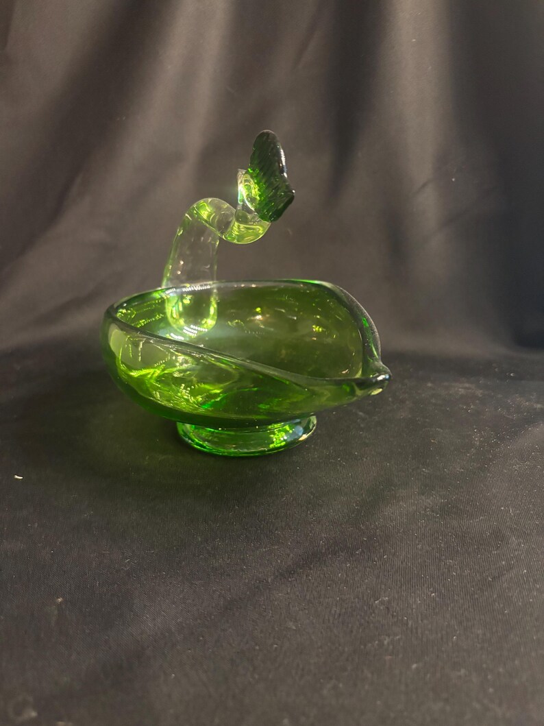 May include: A translucent emerald green glass bowl featuring a curved, swan-like neck handle that extends upward from the rim. The vessel rests on a circular pedestal base and has a smooth, polished finish with a pointed spout on one side.