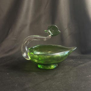 May include: A translucent green glass bowl shaped like a leaf, featuring a clear glass handle that curves upward and terminates in a small, dark green glass leaf accent. The bowl rests on a circular pedestal base and has a pointed, tapered edge.