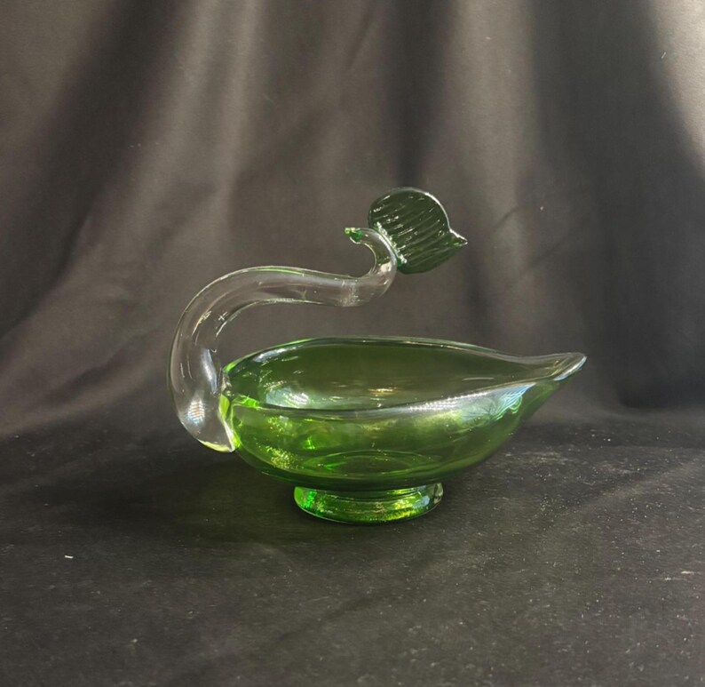 May include: A translucent green glass bowl shaped like a leaf, featuring a clear glass handle that curves upward and terminates in a small, dark green glass leaf accent. The bowl rests on a circular pedestal base and has a pointed, tapered edge.