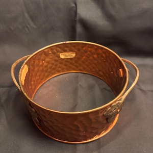 May include: A copper-colored metal bowl with a hammered texture and two handles. The bowl has a circular shape with a decorative leaf design on the side. The interior of the bowl is smooth, and the exterior has a textured finish.