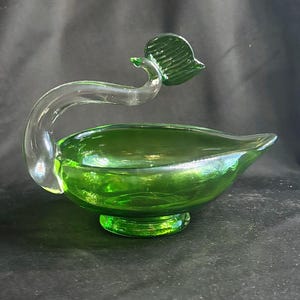 May include: A green glass bowl with a pedestal base and a pointed spout. A clear, curved glass handle extends from the side, topped with a small, textured green glass leaf element. The bowl features a smooth, translucent finish throughout.
