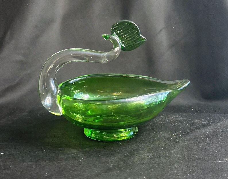 May include: A green glass bowl with a pedestal base and a pointed spout. A clear, curved glass handle extends from the side, topped with a small, textured green glass leaf element. The bowl features a smooth, translucent finish throughout.