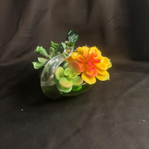 May include: A clear glass swan figurine serving as a planter, filled with artificial succulent plants. The succulents feature shades of green, yellow, and orange. The swan has a long, curved neck and a green glass head, holding the plants within its body.