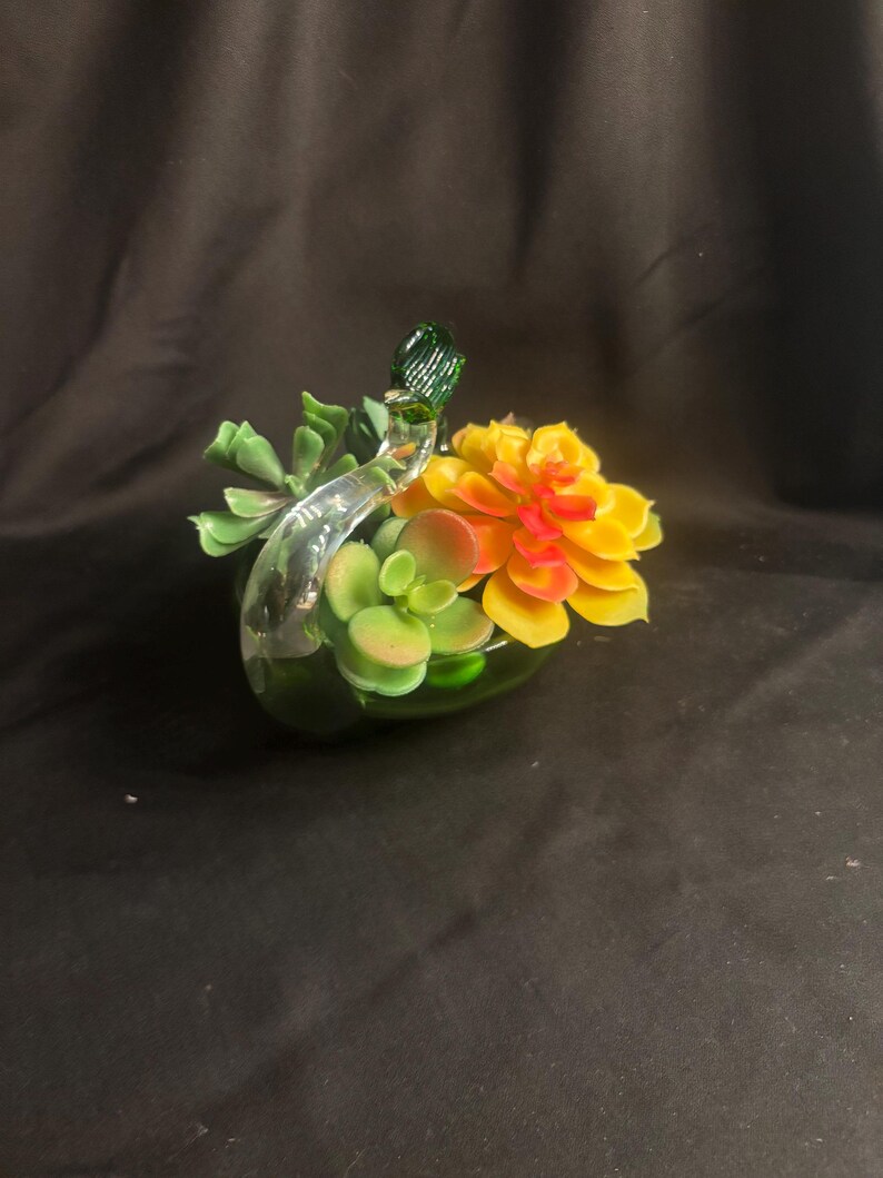 May include: A clear glass swan figurine serving as a planter, filled with artificial succulent plants. The succulents feature shades of green, yellow, and orange. The swan has a long, curved neck and a green glass head, holding the plants within its body.