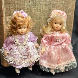 May include: Two miniature porcelain dolls. One doll wears a lavender dress with white lace trim and a matching bow. The other doll is dressed in a pink dress with a matching bonnet and lace trim. Both dolls have blonde hair and are wearing ice skates.