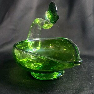 May include: A vibrant green glass bowl featuring a smooth, rounded basin and a pedestal base. A curved, translucent green glass handle extends upward from the rim, terminating in a small, textured, fan-shaped decorative element at the top.