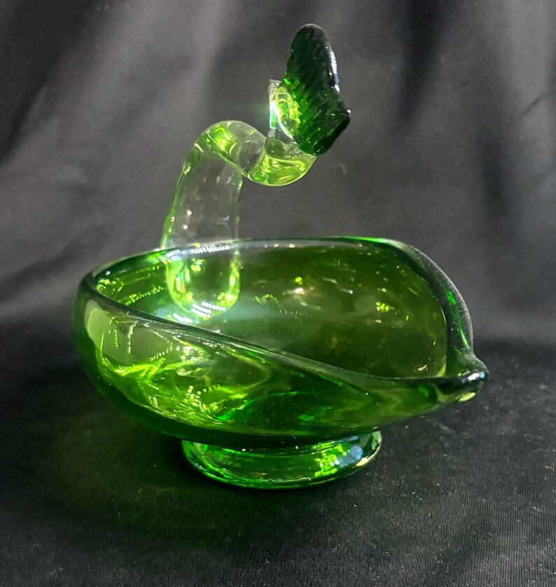 May include: A vibrant green glass bowl featuring a smooth, rounded basin and a pedestal base. A curved, translucent green glass handle extends upward from the rim, terminating in a small, textured, fan-shaped decorative element at the top.