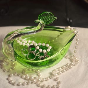 May include: A bright green glass bowl with a curved handle shaped like a leaf. The bowl contains several silver-toned metal rings and is draped with a long strand of white faux pearls that spills over the edges of the glass vessel.