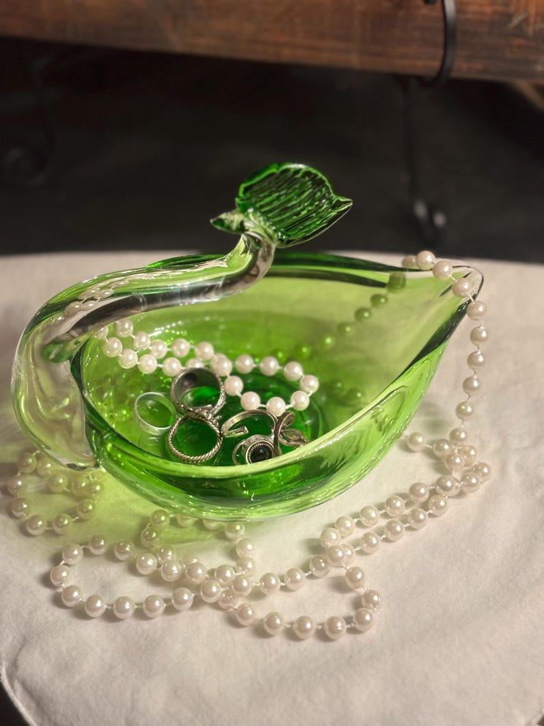May include: A bright green glass bowl with a curved handle shaped like a leaf. The bowl contains several silver-toned metal rings and is draped with a long strand of white faux pearls that spills over the edges of the glass vessel.