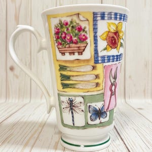 May include: White ceramic mug with a decorative garden theme. The mug features colorful illustrations of flowers, vegetables, and gardening tools. The handle is curved, and the base has a green trim. The mug is approximately 6 inches tall.