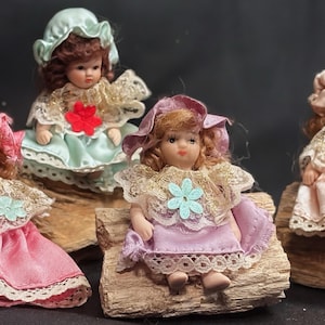 May include: Four porcelain dolls with curly hair, each wearing a bonnet and a dress with lace trim and a small flower accent. The dolls are dressed in pink, light green, lavender, and peach, and are positioned on pieces of textured wood.