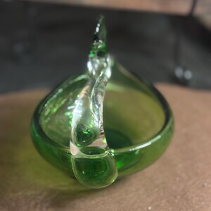 May include: A small, translucent green glass bowl featuring a clear, curved glass handle that arches over the top. The bowl has a smooth, rounded base and a polished finish, showcasing a vibrant emerald green hue throughout the glass structure.