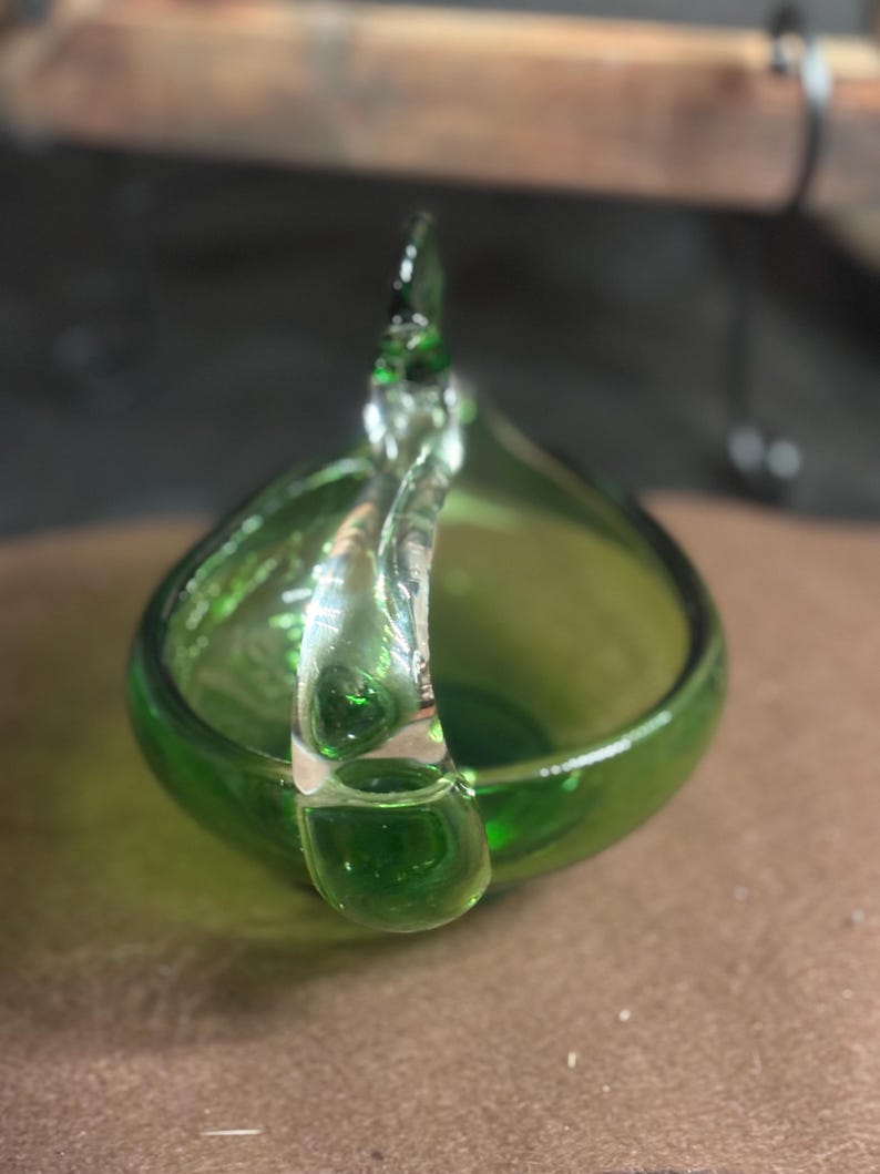 May include: A small, translucent green glass bowl featuring a clear, curved glass handle that arches over the top. The bowl has a smooth, rounded base and a polished finish, showcasing a vibrant emerald green hue throughout the glass structure.