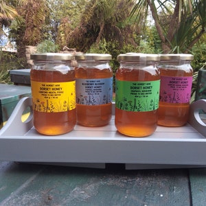 May include: Four glass jars of honey on a grey wooden tray. The jars are labelled with different types of honey, including Canford Heath, Blueberry Blossom, and Dorset Honey. The jars are all 454g/16 oz.