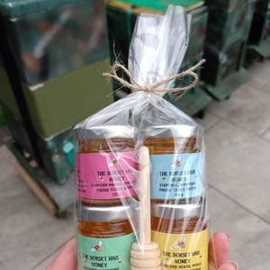 May include: A cellophane bag tied with twine contains four small jars of honey with colourful labels. A wooden honey dipper rests inside. The labels read "The Dorset Hive Honey" and "Proud to Bee British."