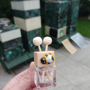 May include: A small glass bottle with a wooden top and a bee design. The bottle has two wooden sticks inside and is clear.
