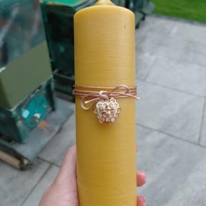May include: A tall, cylindrical beeswax candle in a warm yellow hue. The candle is decorated with a brown cord and a charm with a honeycomb and bee design. The candle is held in a person's hand.