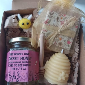 May include: A gift box filled with honey, a beeswax candle shaped like a honeycomb, a wooden honey dipper, and a small felt bee. The honey jar has a pink label that reads "The Dorset Hive Dorset Honey Ford Magna, Wimborne Proud to Bee British 114 g / 4 oz".