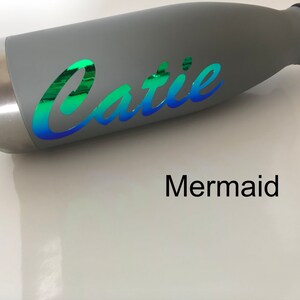 Peut inclure: Bouteille d'eau en acier inoxydable grise avec un autocollant en vinyle iridescent vert, bleu et violet qui dit "Catie" en &eacute;criture cursive. Le mot "Mermaid" est imprim&eacute; en noir sous la bouteille.