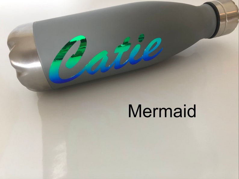 Peut inclure: Bouteille d'eau en acier inoxydable grise avec un autocollant en vinyle iridescent vert, bleu et violet qui dit "Catie" en &eacute;criture cursive. Le mot "Mermaid" est imprim&eacute; en noir sous la bouteille.