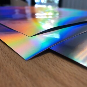 May include: Three sheets of iridescent holographic paper in shades of blue, purple, and green. The paper is stacked on top of each other, with the top sheet folded over the bottom two sheets.