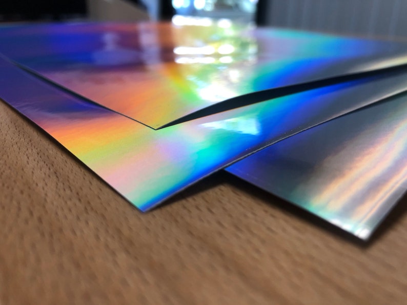 May include: Three sheets of iridescent holographic paper in shades of blue, purple, and green. The paper is stacked on top of each other, with the top sheet folded over the bottom two sheets.