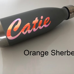 Peut inclure: Une bouteille d'eau en acier inoxydable grise avec un autocollant en vinyle iridescent rose et orange qui dit "Catie" en &eacute;criture cursive. La bouteille est sur une surface blanche. Orange Sorbet.