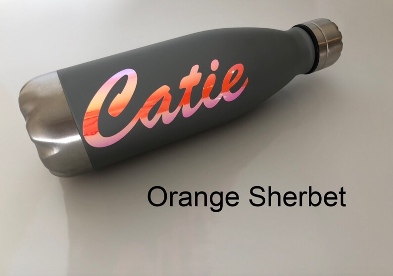 Peut inclure: Une bouteille d'eau en acier inoxydable grise avec un autocollant en vinyle iridescent rose et orange qui dit "Catie" en &eacute;criture cursive. La bouteille est sur une surface blanche. Orange Sorbet.