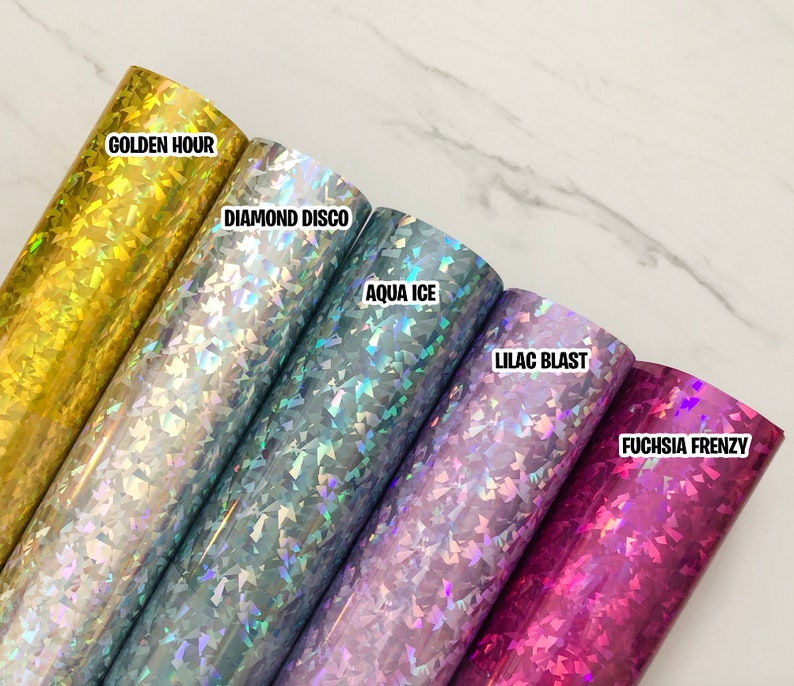 May include: Five rolls of iridescent vinyl with holographic glitter in different colors. The colors are gold, silver, blue, purple, and pink. The rolls are labeled "Golden Hour", "Diamond Disco", "Aqua Ice", "Lilac Blast", and "Fuchsia Frenzy".
