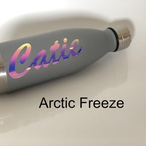 Peut inclure: Une bouteille d'eau en acier inoxydable grise avec un autocollant holographique qui dit "Catie". La bouteille est sur une surface blanche et le texte "Arctic Freeze" est en dessous de la bouteille.