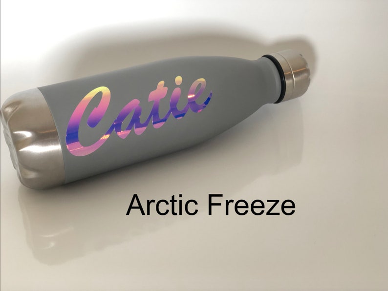 Peut inclure: Une bouteille d'eau en acier inoxydable grise avec un autocollant holographique qui dit "Catie". La bouteille est sur une surface blanche et le texte "Arctic Freeze" est en dessous de la bouteille.