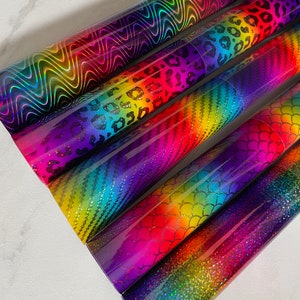 May include: Five rolls of iridescent vinyl with different patterns, including a rainbow stripe, a mermaid scale, a leopard print, and a wavy line pattern. The vinyl is holographic and has a rainbow sheen.