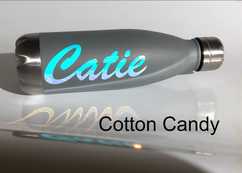 Peut inclure: Bouteille d'eau en acier inoxydable grise avec un autocollant en vinyle iridescent bleu turquoise qui dit "Catie" en &eacute;criture cursive. La bouteille est pos&eacute;e sur une surface blanche avec les mots "Cotton Candy" imprim&eacute;s en dessous.