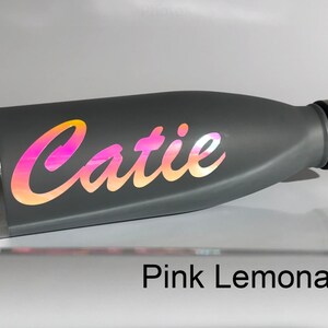 Peut inclure: Une bouteille d'eau en acier inoxydable grise avec un autocollant en vinyle iridescent rose et orange qui dit "Catie". La bouteille est pos&eacute;e sur une surface blanche. Le texte "Pink Lemonade" est visible sous la bouteille.
