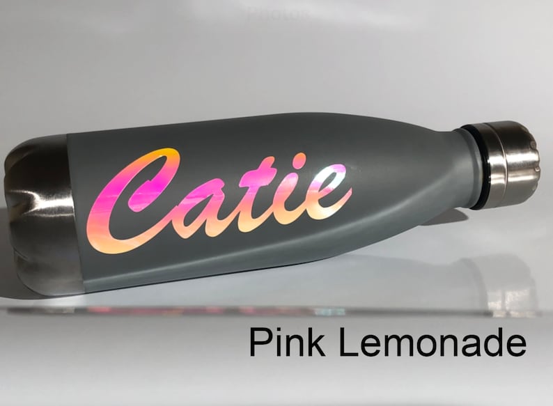 Peut inclure: Une bouteille d'eau en acier inoxydable grise avec un autocollant en vinyle iridescent rose et orange qui dit "Catie". La bouteille est pos&eacute;e sur une surface blanche. Le texte "Pink Lemonade" est visible sous la bouteille.