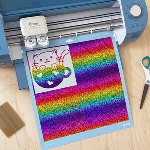 May include: A Cricut cutting machine with a rainbow glitter vinyl sheet featuring a white silhouette of a cat in a mug. The cat has a cute face and the mug has hearts on it.