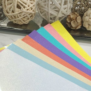 K&ouml;nnte beinhalten: Ein Stapel aus sieben Glitzerpapierb&ouml;gen in verschiedenen Pastellfarben, darunter Wei&szlig;, Hellblau, Creme, Pfirsich, Lavendel, T&uuml;rkis und Gelb.