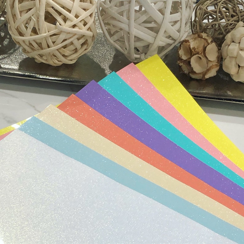 K&ouml;nnte beinhalten: Ein Stapel aus sieben Glitzerpapierb&ouml;gen in verschiedenen Pastellfarben, darunter Wei&szlig;, Hellblau, Creme, Pfirsich, Lavendel, T&uuml;rkis und Gelb.