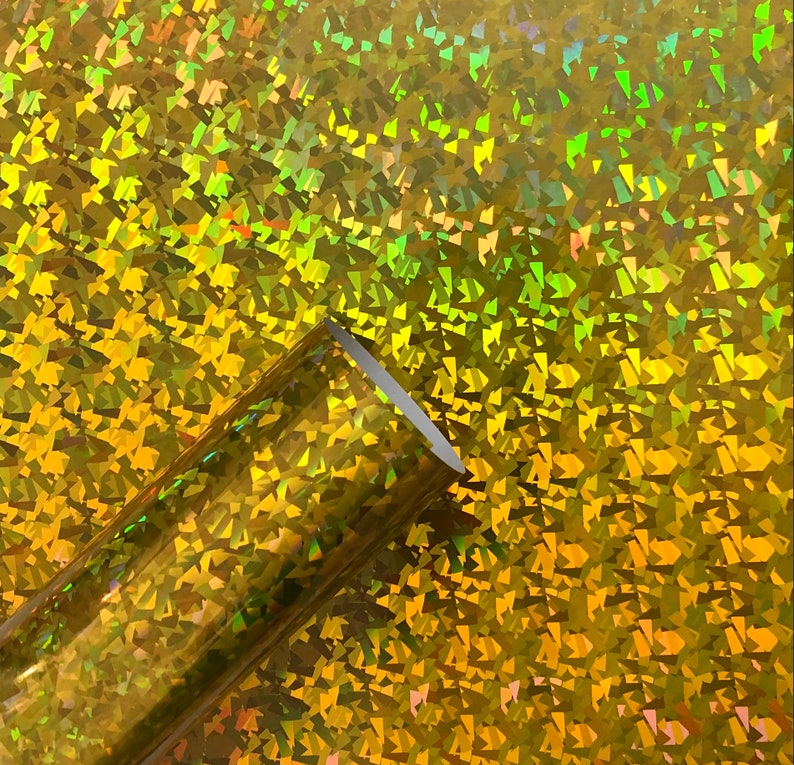 May include: A roll of iridescent gold holographic vinyl with a textured, geometric pattern. The vinyl is shiny and reflective.