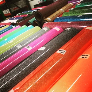 May include: An assortment of glitter vinyl rolls in shades like neon orange, pink, and black. Each roll is labelled with its colour name. The rolls are displayed on a table, presenting a range of crafting materials.