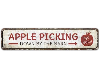 May include: A white metal sign with red text that reads "Apple Picking - Down by the Barn" with a red arrow pointing to the right. A red apple with a green stem and leaf is on the right side of the sign with the text "5¢ each".