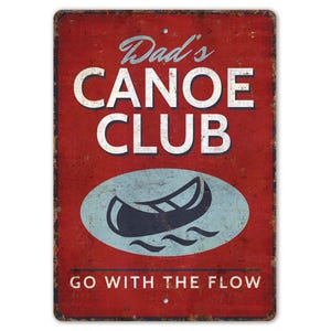 May include: A rectangular, vintage-style metal sign with a distressed red background. The sign features the text "Dad's CANOE CLUB" in white, with a canoe graphic in a blue oval. The phrase "GO WITH THE FLOW" is at the bottom.
