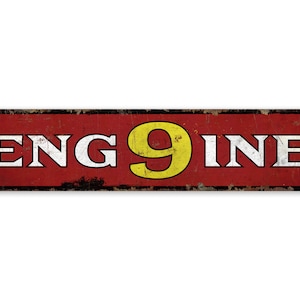 May include: A rectangular, vintage-style metal sign with the word "ENGINE" in white capital letters and a yellow number "9" in the middle. The word "INE" is also in white. The sign has a red background with a distressed, rusty appearance.