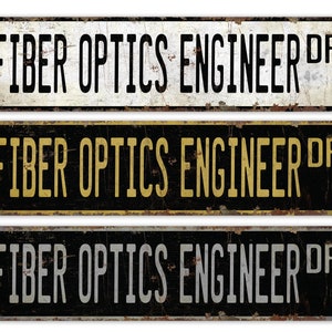 Op de afbeelding: Drie vintage-stijl metalen borden met de tekst "Fiber Optics Engineer Dr." in zwarte letters op een witte, zwarte en grijze achtergrond.