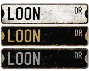 May include: Three vintage-style metal street signs. The signs are rectangular with rounded corners and a distressed finish. Each sign reads "LOON DR" in bold, uppercase letters. The signs have a rustic, aged appearance.