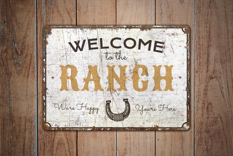 May include: White metal sign with distressed paint and rusty edges. The sign reads "Welcome to the Ranch" in black and gold lettering. A horseshoe is in the centre of the sign with the text "We're Happy You're Here" below.