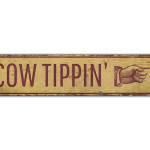 May include: A rustic metal sign with a yellow background and red text that reads "COW TIPPIN'" with a red pointing hand graphic.